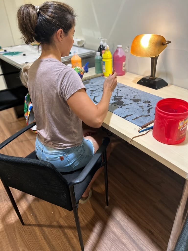 A parent painting at a studio desk under a warm lamp, with paint bottles and brushes nearby