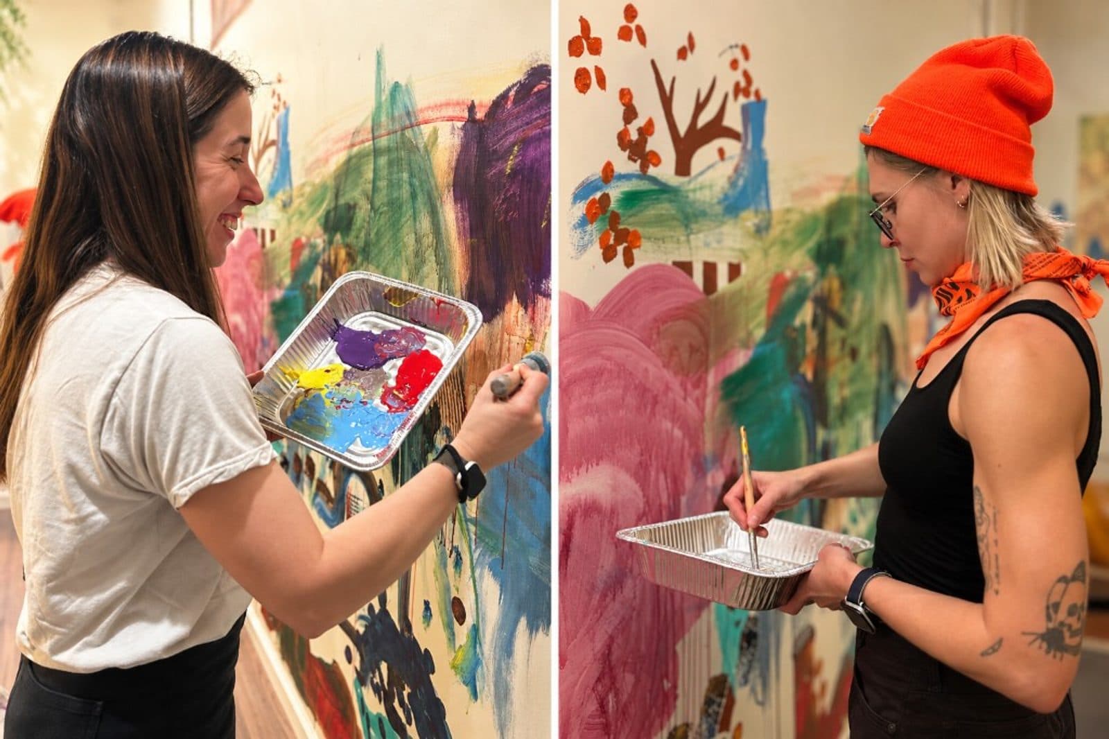 Two adults painting on the studio's shared wall, one laughing with a palette of bright paint, the other quietly loading her brush