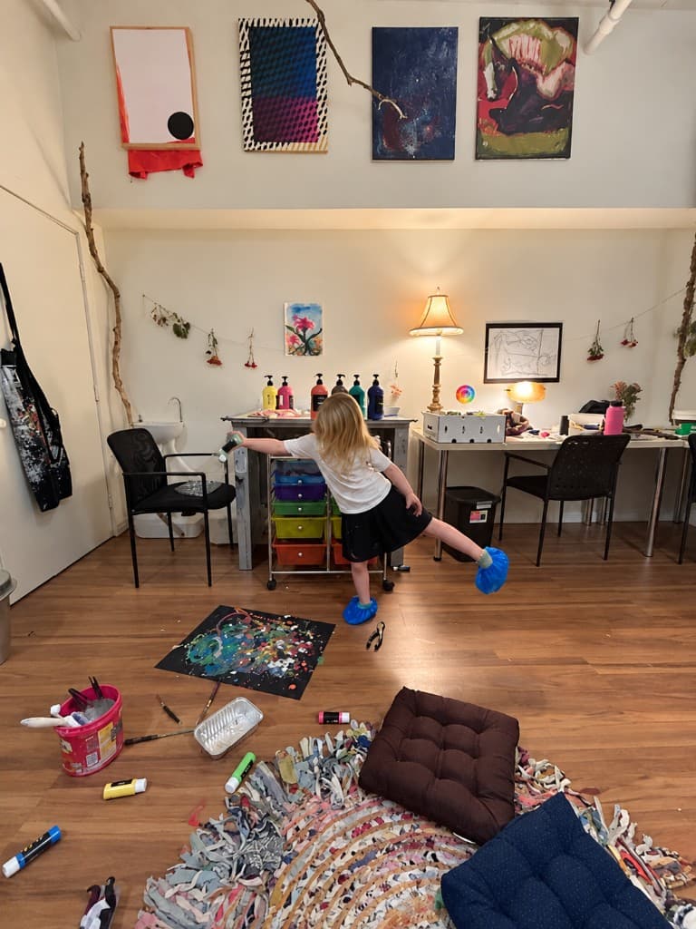 A girl balancing on one leg in blue shoe covers, reaching toward a rolling cart of paint in the studio