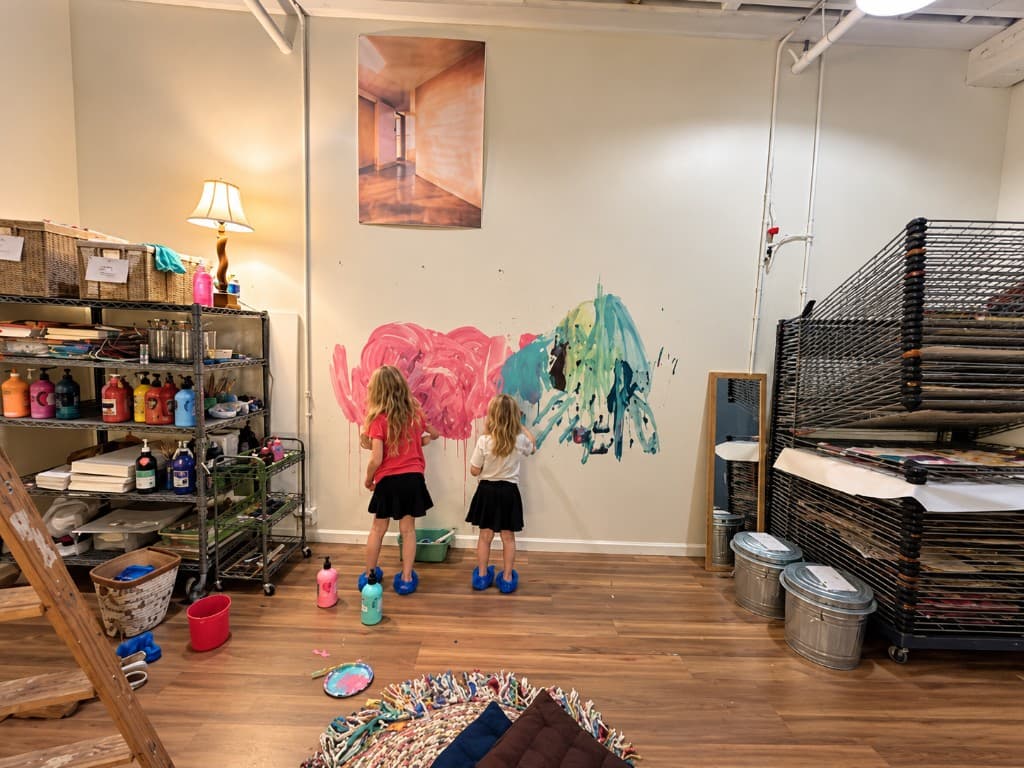 Two children painting on a studio wall, with supply shelves, drying racks, and materials visible across the space